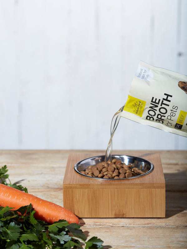 Pouring chicken bone broth from a pouch into a pet food bowl with fresh carrot and parsley nearby
